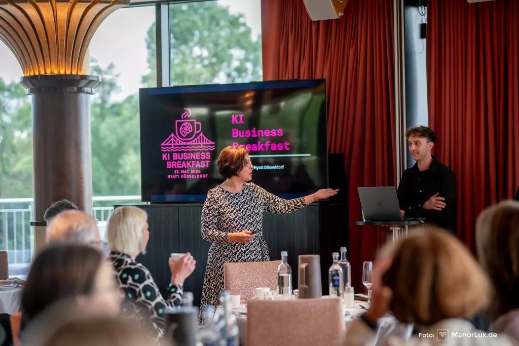 KI Business Breakfast. Speaker Niklas und Moderatorin Martha stehen in einem Raum voller Gäste an kleinen Tischgruppen im Hyatt Düsseldorf
